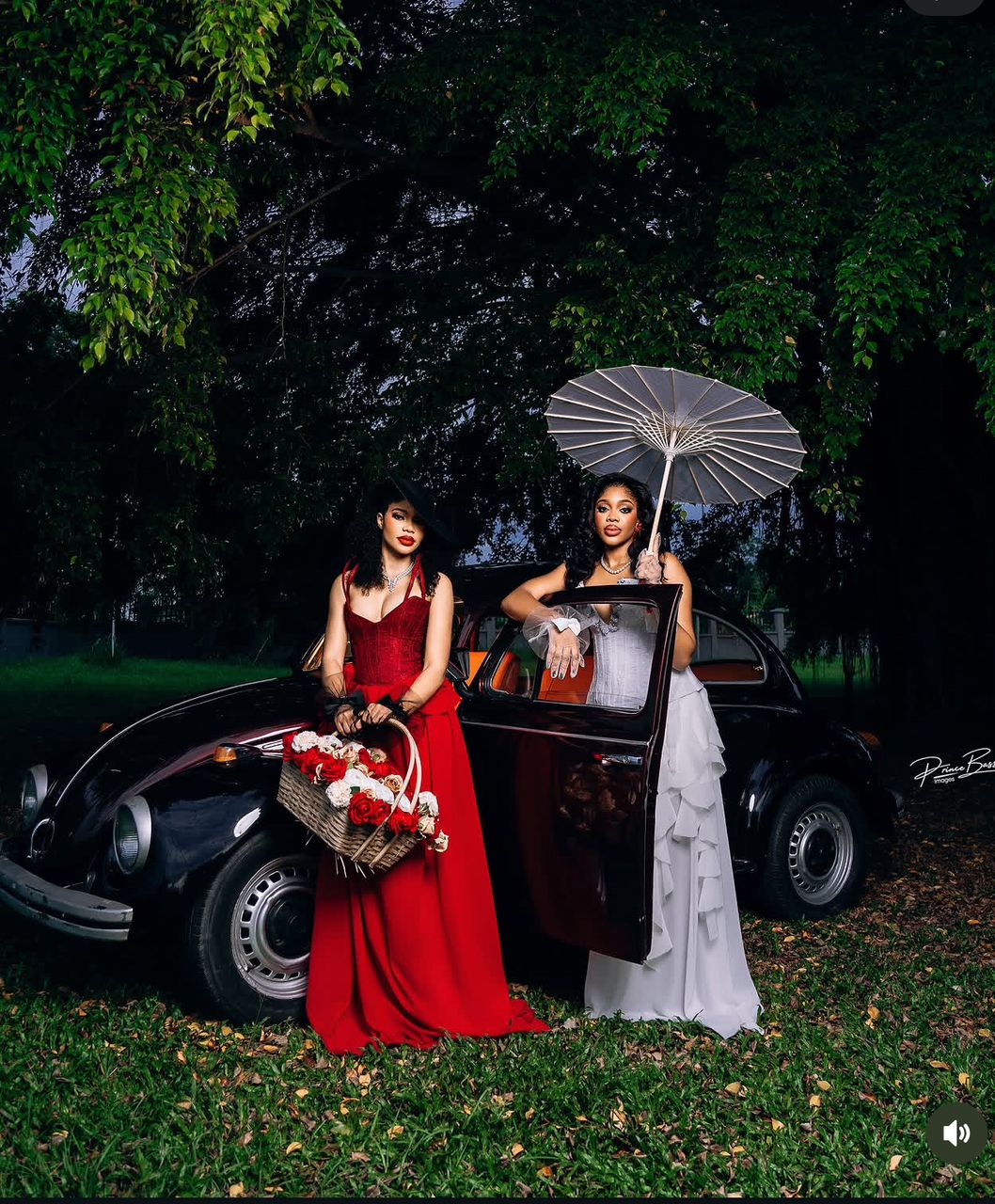 Two women in evening gowns with statement jewelry beside a vintage car